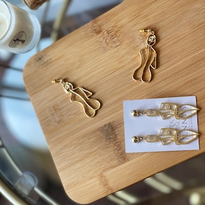 以下が含まれることがあります： 抽象的な人間の図形が描かれた金色のイヤリングが3組。イヤリングは木製の表面に置かれています。