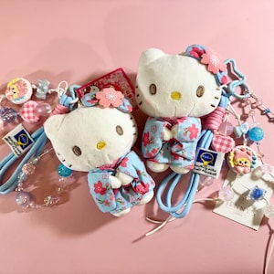 May include: Two Hello Kitty plushies wearing floral patterned kimonos, with matching hair ties and beaded accessories. The plushies are white with brown eyes and yellow noses. The background is a solid pink.