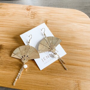 May include: A pair of gold-toned earrings featuring a fan-shaped design with intricate details. Each earring has a small white pearl charm and a delicate gold chain dangling from the bottom.