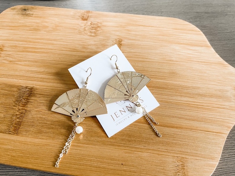 May include: A pair of gold-toned earrings featuring a fan-shaped design with intricate details. Each earring has a small white pearl charm and a delicate gold chain dangling from the bottom.