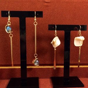 May include: Gold-toned dangle earrings on display. One pair features a blue crystal and a long bar, the other has a white, irregular-shaped stone and a long bar. The earrings are presented on black stands against a red background.