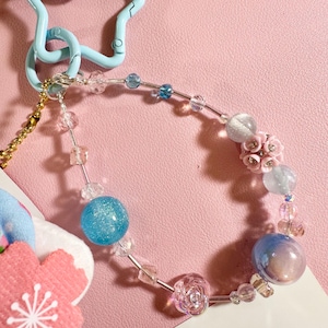 May include: A beaded charm bracelet with a light blue star-shaped clasp. The bracelet features clear, blue, and pink beads, including a large glittery blue sphere and a pink flower. A small pink and blue flower charm is also attached.