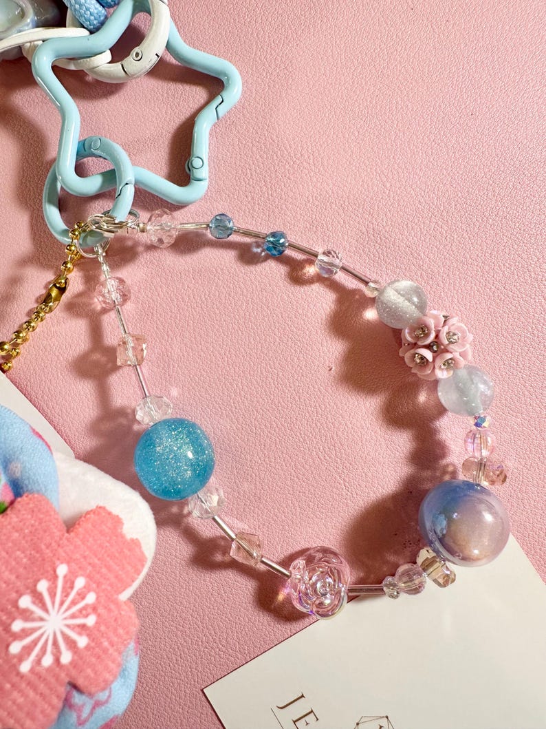 May include: A beaded charm bracelet with a light blue star-shaped clasp. The bracelet features clear, blue, and pink beads, including a large glittery blue sphere and a pink flower. A small pink and blue flower charm is also attached.