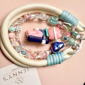 May include: A handmade dog leash with a white rope, light blue accents, and a variety of colorful beads. A small figurine is in the center. The leash is on a pink surface. The brand name "JENNY'S CORNER" is visible.