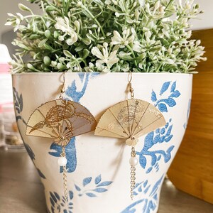May include: A pair of gold fan-shaped earrings with a pearl dangle. The earrings are hanging from a white planter with blue floral designs.