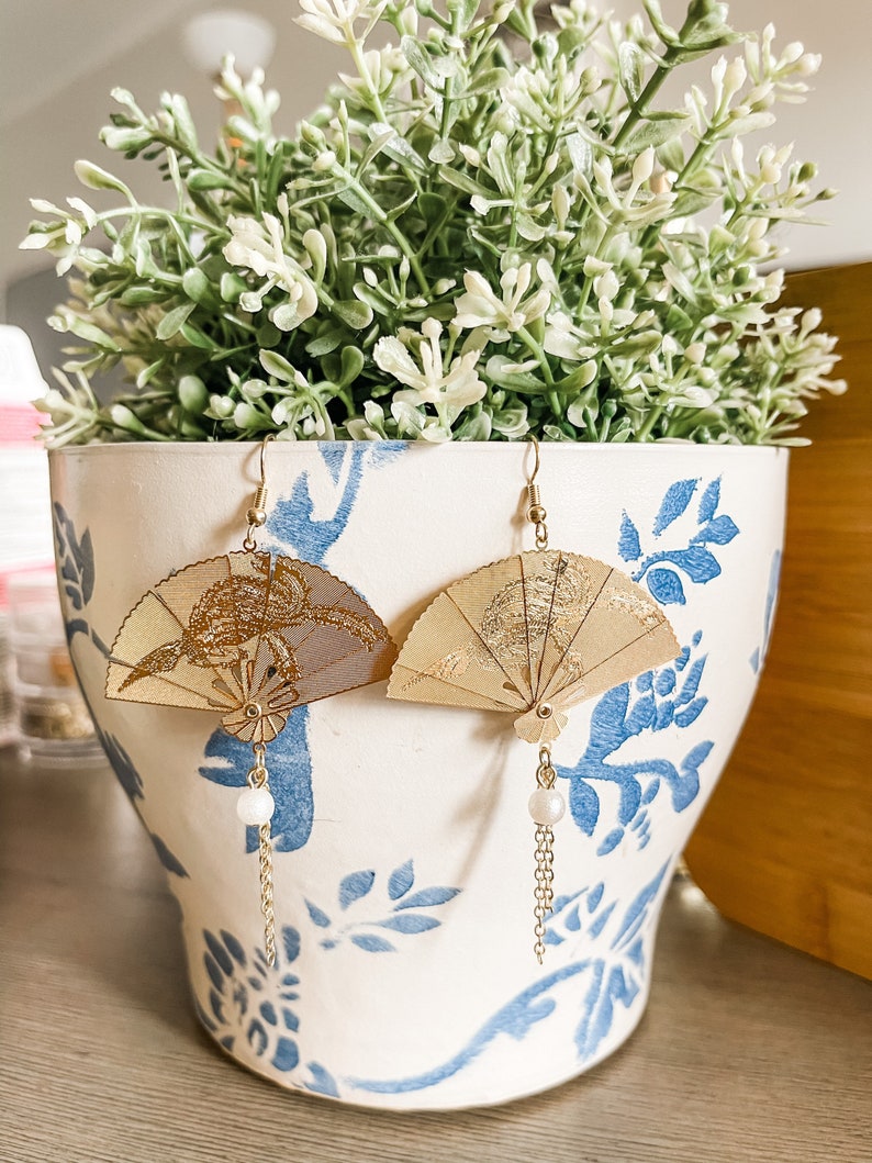 May include: A pair of gold fan-shaped earrings with a pearl dangle. The earrings are hanging from a white planter with blue floral designs.