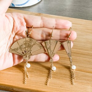 May include: A pair of gold-tone earrings featuring a fan-shaped design with a pearl accent and a dangling chain. The earrings are displayed on a wooden surface.