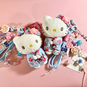 May include: Two Hello Kitty plushies wearing floral kimonos, with matching blue straps and beaded accents. The plushies have pink flower hair accessories and are set against a pink background. The accessories include small charms and the text "JENNY'S".
