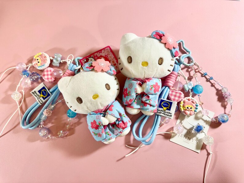May include: Two Hello Kitty plushies wearing floral kimonos, with matching blue straps and beaded accents. The plushies have pink flower hair accessories and are set against a pink background. The accessories include small charms and the text "JENNY'S".