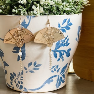 May include: A pair of gold fan-shaped earrings with a pearl and chain dangle. The earrings are displayed on a white ceramic planter with blue floral designs.