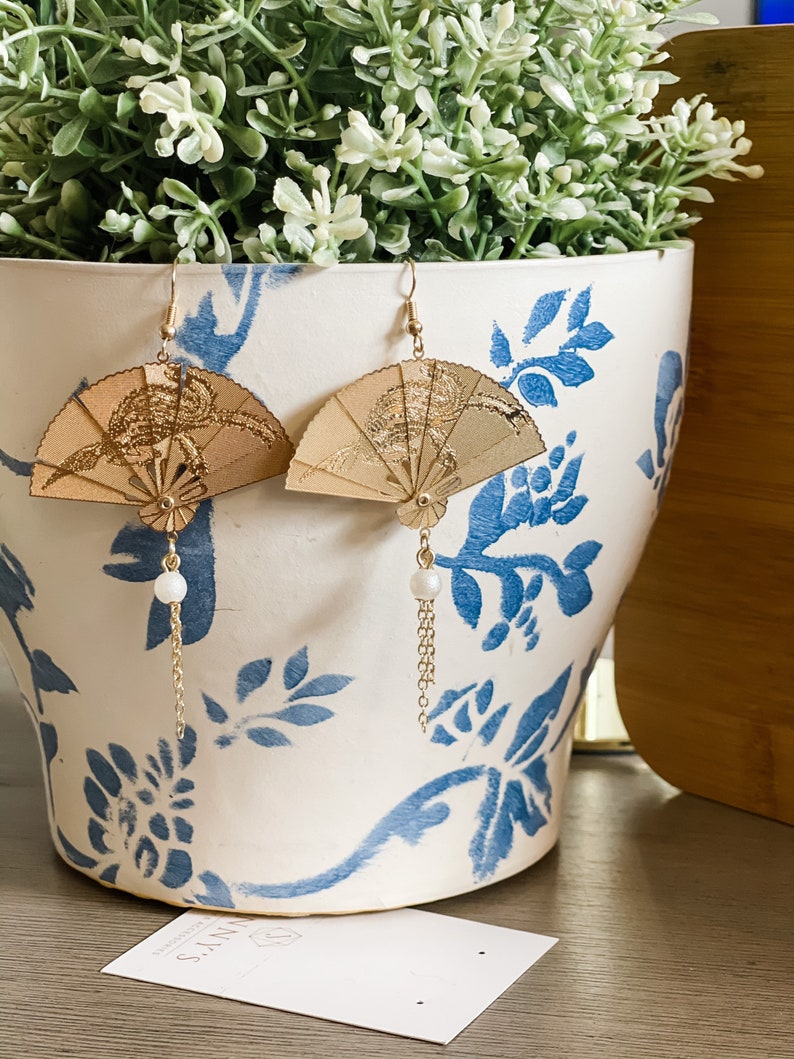 May include: A pair of gold fan-shaped earrings with a pearl and chain dangle. The earrings are displayed on a white ceramic planter with blue floral designs.
