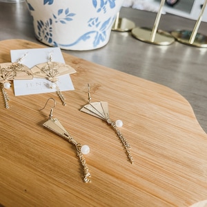 May include: Two pairs of gold fan-shaped earrings with pearl accents and dangling chains. The earrings are displayed on a wooden surface.