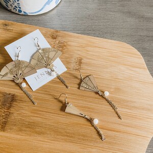 May include: Three pairs of gold-toned earrings with a fan-shaped design. The fans are made of paper and have a gold-toned finish. The earrings are hanging from a gold-toned chain with a pearl bead at the end.