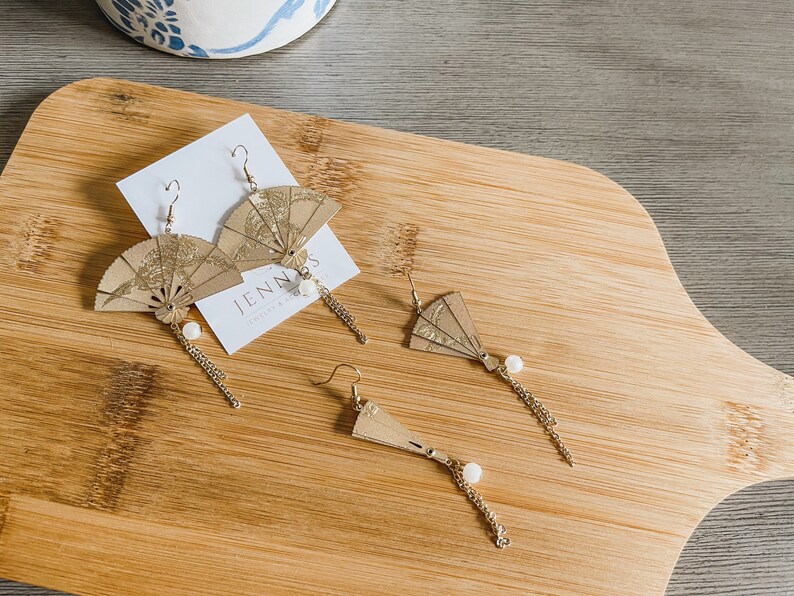 May include: Three pairs of gold-toned earrings with a fan-shaped design. The fans are made of paper and have a gold-toned finish. The earrings are hanging from a gold-toned chain with a pearl bead at the end.