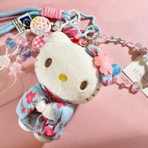 May include: A plush Hello Kitty doll wearing a floral kimono, adorned with a pink flower and blue accents. The doll is attached to a light blue lanyard with decorative beads and charms. A tag with the name "JENNY'S" is visible.