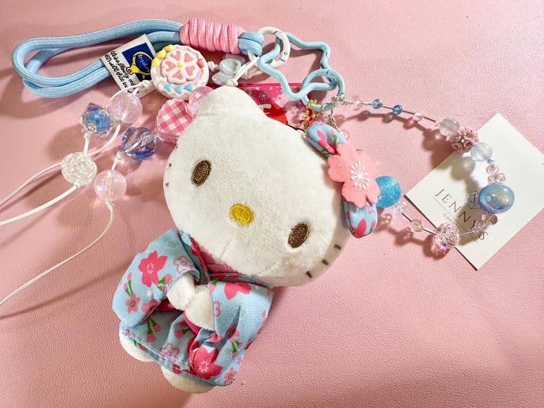 May include: A plush Hello Kitty doll wearing a floral kimono, adorned with a pink flower and blue accents. The doll is attached to a light blue lanyard with decorative beads and charms. A tag with the name "JENNY'S" is visible.