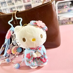 May include: A white plush Hello Kitty keychain wearing a blue floral kimono and a pink flower hair accessory. The keychain is attached to a brown purse with a light blue and pink beaded charm. The background is a pink surface.