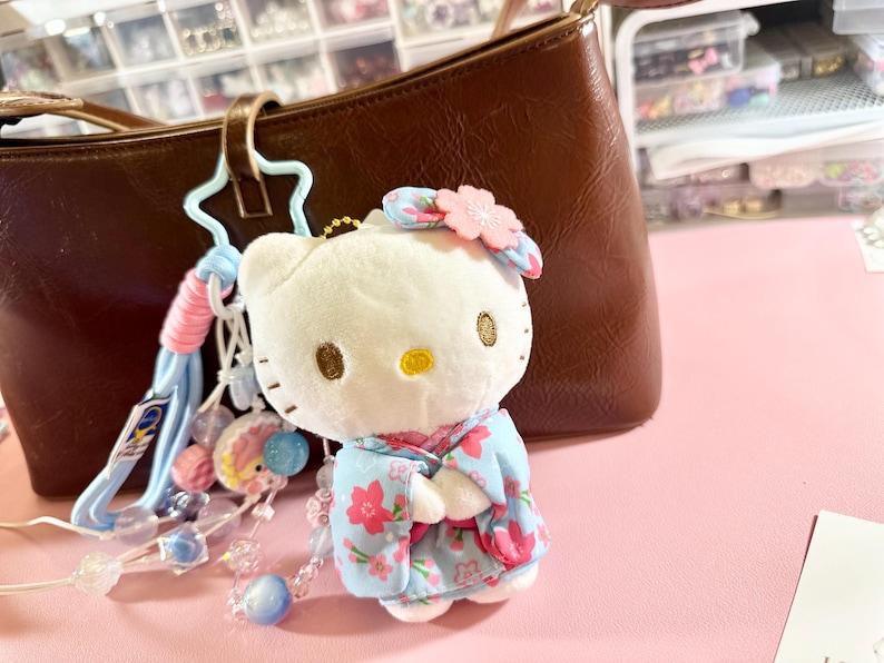 May include: A white plush Hello Kitty keychain wearing a blue floral kimono and a pink flower hair accessory. The keychain is attached to a brown purse with a light blue and pink beaded charm. The background is a pink surface.