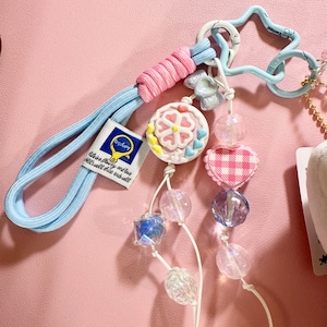 May include: A light blue keychain with a pink and blue cord, a star-shaped clasp, and a charm with a flower design. The keychain also features a series of clear and iridescent beads, a pink gingham heart, and a tag that reads "life is a play..."