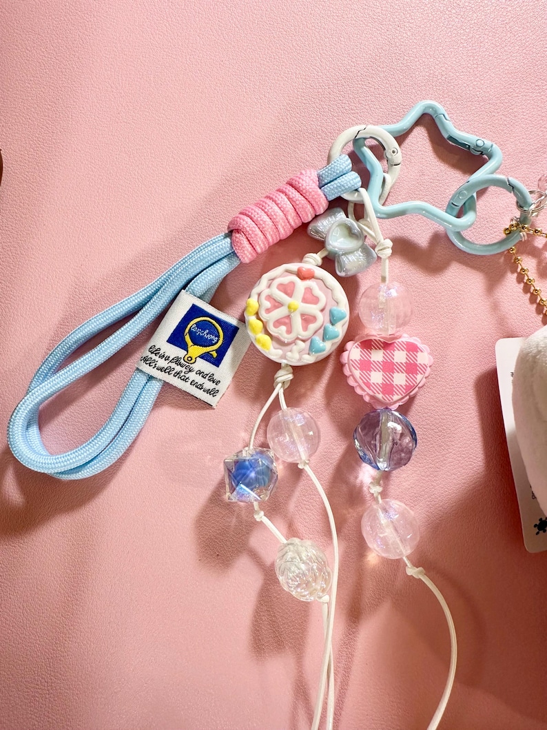 May include: A light blue keychain with a pink and blue cord, a star-shaped clasp, and a charm with a flower design. The keychain also features a series of clear and iridescent beads, a pink gingham heart, and a tag that reads "life is a play..."