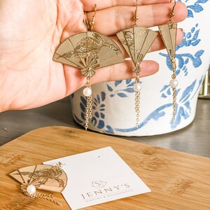 May include: Gold tone earrings with a fan-shaped design and a pearl dangle. The earrings are on a wooden surface with a white card that says "Jenny's Jewelry & Accessories".