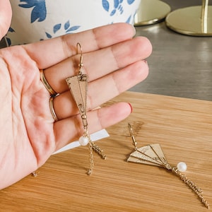 May include: A pair of gold earrings with a pearl and a fan-shaped charm. The fan is made of a beige material and has a gold trim.