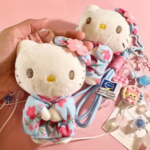 May include: Two plush white cat-shaped dolls with embroidered facial features and wearing light blue floral patterned outfits. One doll is a keychain. The dolls have pink flower accents and are attached to a blue lanyard with decorative beads. The tag reads "JENNY'S CORNER".