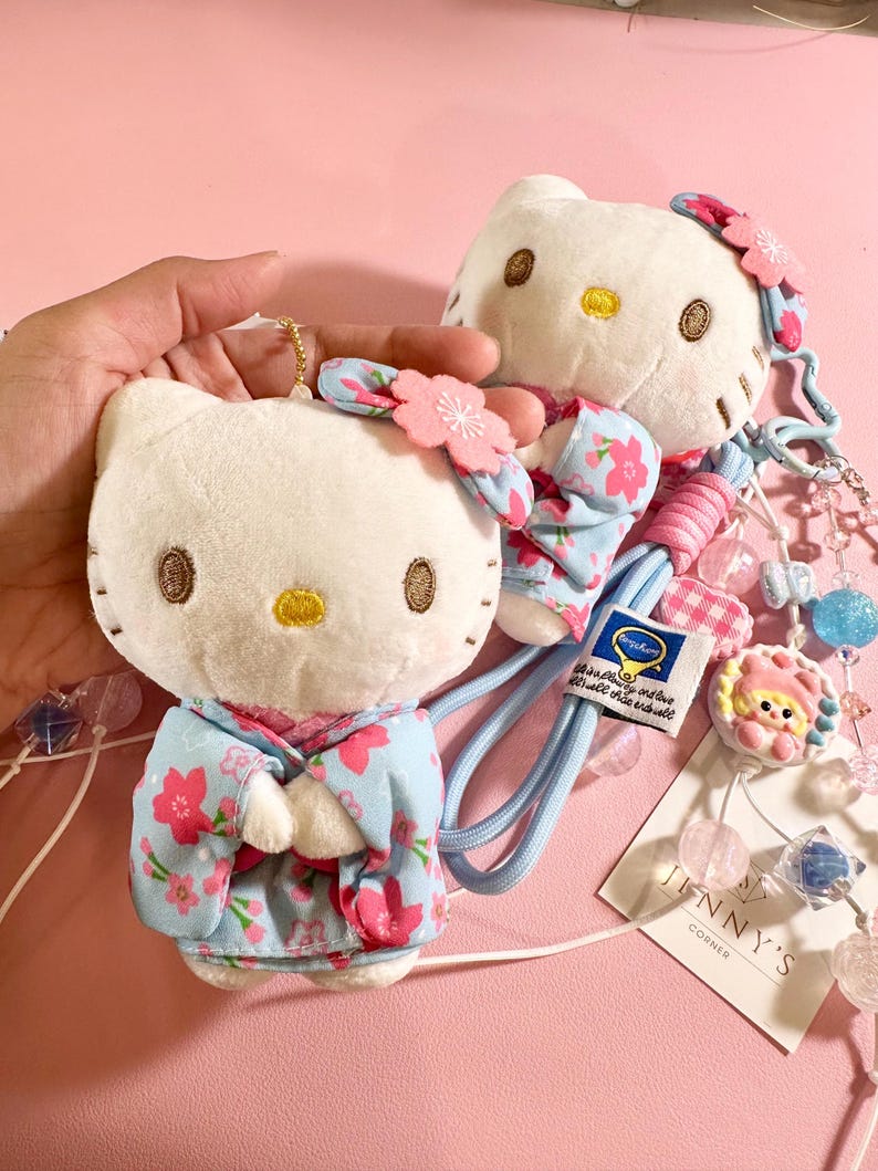 May include: Two plush white cat-shaped dolls with embroidered facial features and wearing light blue floral patterned outfits. One doll is a keychain. The dolls have pink flower accents and are attached to a blue lanyard with decorative beads. The tag reads "JENNY'S CORNER".