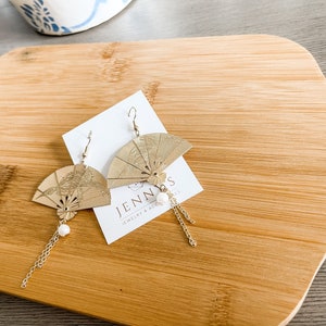 May include: Gold fan-shaped earrings with a pearl accent and a gold chain dangle. The earrings are on a white card with the text "Jenn's Jewelry & Accessories".