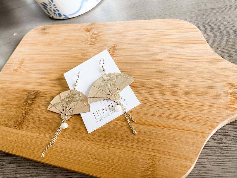 May include: Gold fan-shaped earrings with a pearl accent and a gold chain dangle. The earrings are on a white card with the text "Jenn's Jewelry & Accessories".