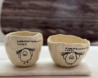 Può includere: Due ciotole in ceramica color bianco sporco, modellate a mano, con bordi irregolari e ondulati e texture maculata. Ogni ciotola presenta un disegno a linee nere di una figura con capelli lunghi e il testo "sisterrrrrrrrrrrrrrrrr" scritto sopra.