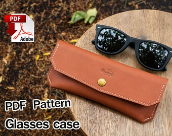 May include: A brown leather glasses case with white stitching and a brass button. The case is closed and has the text "Wilderness" embossed on the front. A pair of black sunglasses is lying on top of the case. The text "PDF Pattern Glasses case" is written below the case. The text "PDF" is written in red on a white download icon with the Adobe logo.
