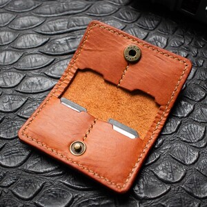 May include: A tan leather card holder featuring visible stitching, two internal slots, and a brass snap button closure. The leather has a smooth finish with a natural grain texture. The text AITA CRAFT Handcrafted leather goods is visible.