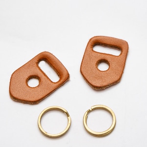 Puede incluir: Dos llaveros de cuero marrón con un anillo de metal unido a cada uno. Los llaveros tienen una forma rectangular con un agujero en el centro y un agujero más pequeño en la parte superior.