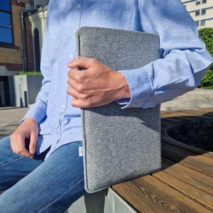 Puede incluir: Una funda para portátil de fieltro gris con un logotipo blanco que dice "TOKU" en la parte delantera.