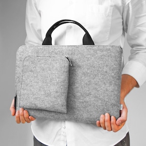 Puede incluir: Una bolsa para portátil de fieltro gris con asa negra y cierre de cremallera. La bolsa tiene un bolsillo más pequeño en la parte delantera.