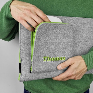 Könnte beinhalten: Eine graue Filz-Laptoptasche mit einem grünen Reißverschluss und dem Namen "Thomas" in grün auf der Vorderseite gestickt.