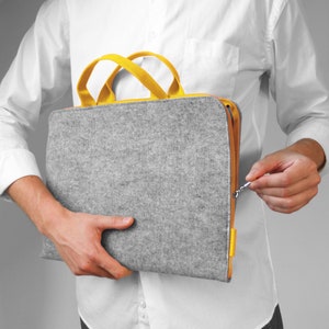 May include: A grey felt laptop bag with yellow handles and a zip closure. The bag is being held by a person.