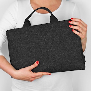 Könnte beinhalten: Eine dunkelgraue Filz-Laptoptasche mit schwarzem Griff. Die Tasche wird von einer Person mit rotem Nagellack gehalten.