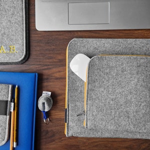 Felt laptop case with pocket for wires sewn on the main part of the sleeve. 15 inch laptop lying on the desk next to sleeve. Case with Yellow zipper. Felt mouse pad with initials next to the laptop.