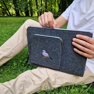 Man holds dark grey felt laptop case with bird embroided on the front. Laptop sleeve has white zipper and front pocket for wires. On the pocket there is an embroidery of long-tailed tit. Cover takes 13 inch laptop.