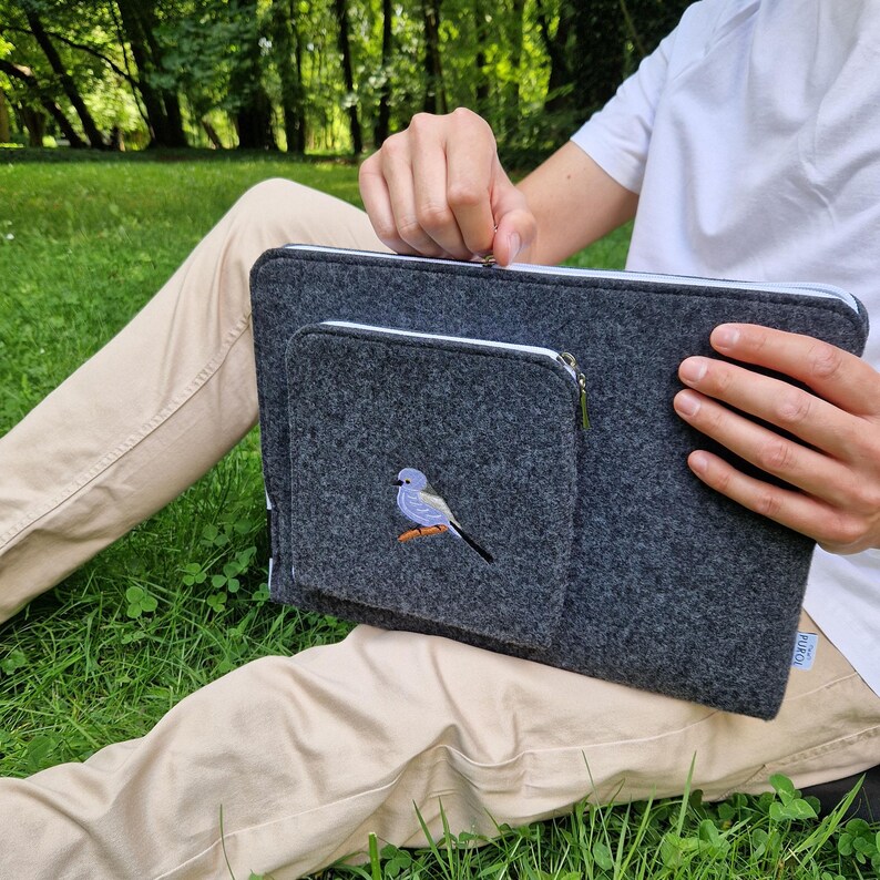 Man holds dark grey felt laptop case with bird embroided on the front. Laptop sleeve has white zipper and front pocket for wires. On the pocket there is an embroidery of long-tailed tit. Cover takes 13 inch laptop.