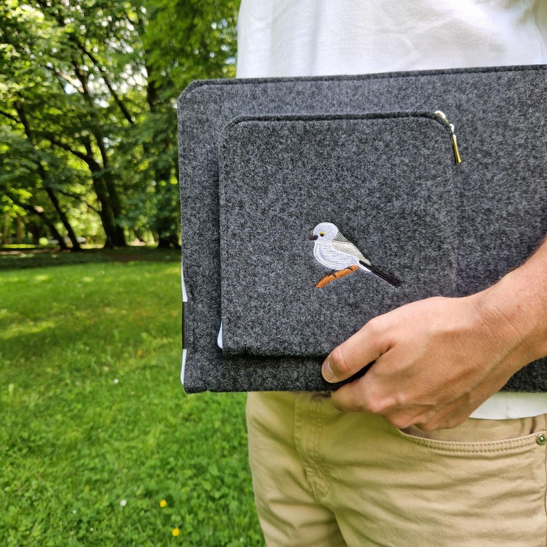 Man holds dark grey felt laptop case with bird embroided on the front. Laptop sleeve has white zipper and front pocket for wires. On the pocket there is an embroidery of long-tailed tit. Cover takes 13 inch laptop.
