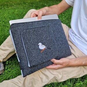 Man holds dark grey felt laptop case with bird embroided on the front. Laptop sleeve has white zipper and front pocket for wires. On the pocket there is an embroidery of long-tailed tit. Cover takes 13 inch laptop.