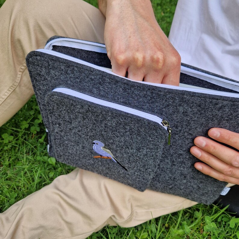 Man holds dark grey felt laptop case with bird embroided on the front. Laptop sleeve has white zipper and front pocket for wires. On the pocket there is an embroidery of long-tailed tit. Cover takes 13 inch laptop.