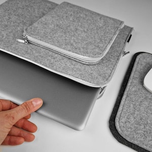 Silver 15 inch laptop half out of the light grey felt sleeve. Lying on the table next to mouse pad.