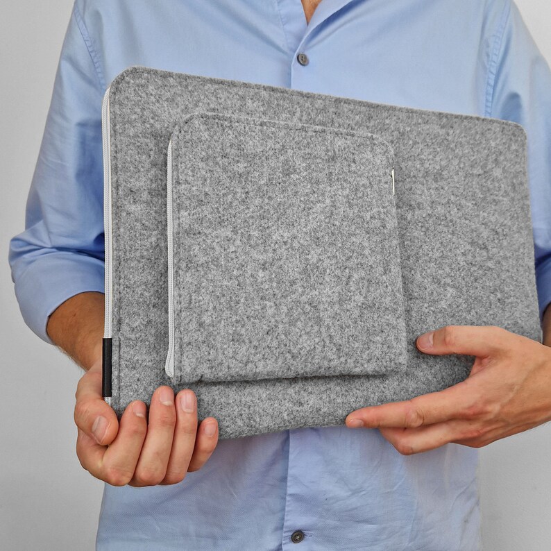 Man holding light grey felt laptop sleeve. Front pocket sewn on the main part of the sleeve. White zipper in the sleeve and in the pocket.