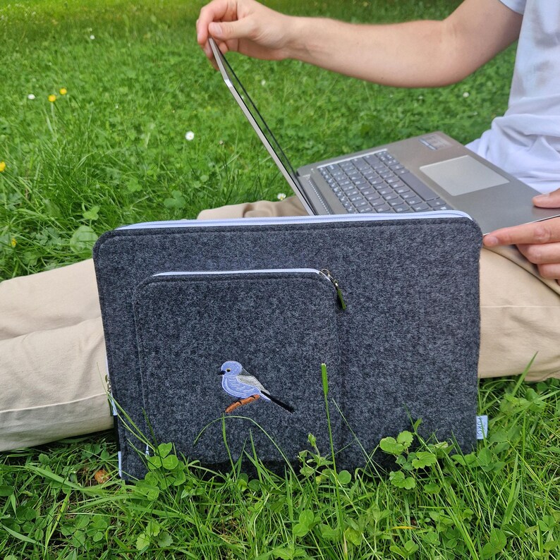 Man holds dark grey felt laptop case with bird embroided on the front. Laptop sleeve has white zipper and front pocket for wires. On the pocket there is an embroidery of long-tailed tit. Cover takes 13 inch laptop.