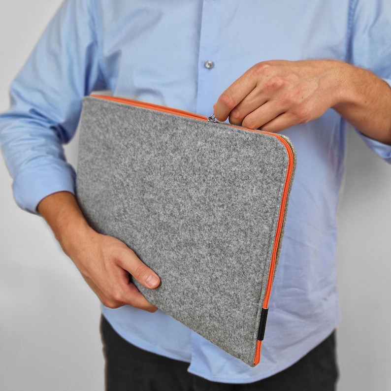 Light grey felt laptop sleeve with orange zipper. Man holding a felt case with 15 inch laptop inside. Orangezipper opens on the corner of the sleeve.