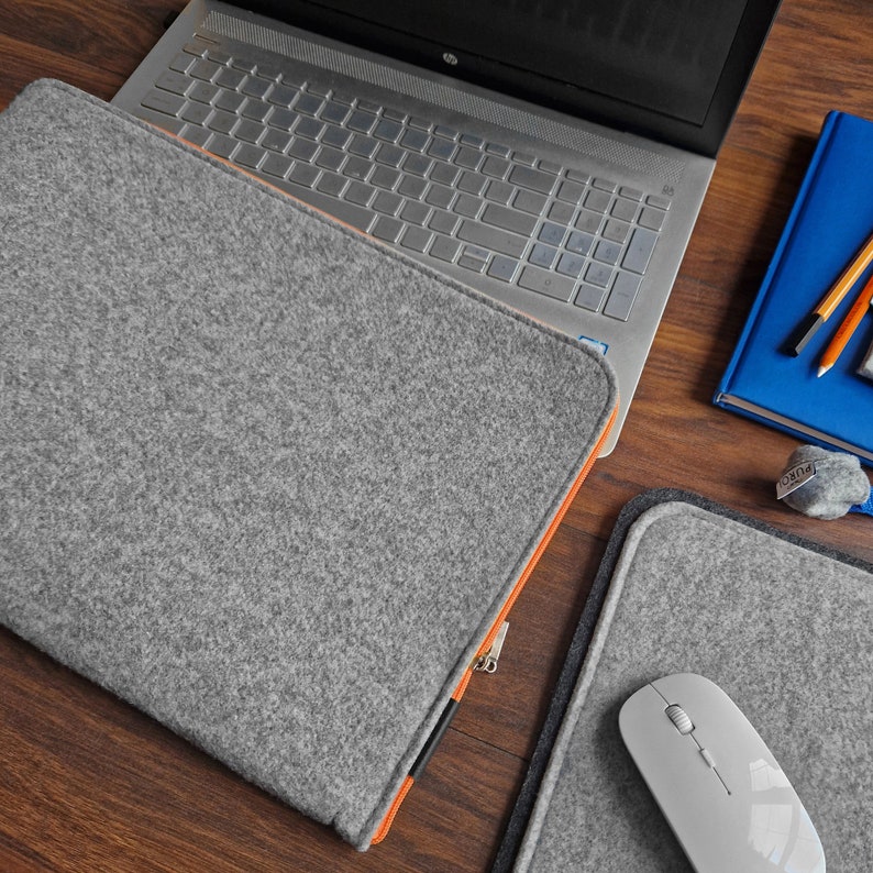 Light grey felt laptop sleeve with orange zipper. 15 inch laptop lying next to felt mouse pad and felt key ring.